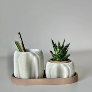 May include: A white, ribbed desk organizer set with a matching tray. One container holds pens and a small plant sits in the other. The set is a neutral color and would be a great addition to any desk or office space.