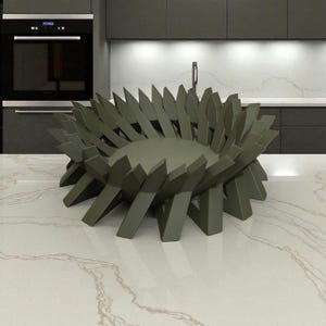May include: A modern, olive green decorative bowl with a unique, spiky design. The bowl is made of a solid material and sits on a white countertop with subtle veining. The kitchen setting includes dark gray cabinets and a built-in oven.