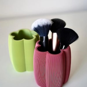 May include: A close-up shot of two decorative containers, one green and one pink, each holding makeup brushes. The pink container holds three brushes with black and white bristles. The containers have a ribbed design and a unique, flower-like shape.