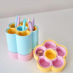 May include: A pastel-colored desk organizer set. The set includes a flower-shaped container and a pen holder. The pen holder is light blue with yellow and pink accents, and holds pens. The flower-shaped container is yellow and pink and holds paper clips.