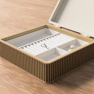 May include: Open jewelry box with a light brown exterior and a cream-colored interior. The box is divided into compartments with white velvet-lined slots for rings and necklaces. A silver ring and two pairs of earrings are visible inside.