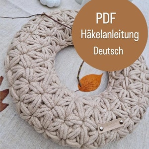 May include: A beige, handmade crochet wreath with a floral pattern. The wreath is circular and features a leather tag. The image includes a small white pumpkin, candles, and fall foliage. Text on the image reads "PDF Häkelanleitung Deutsch."