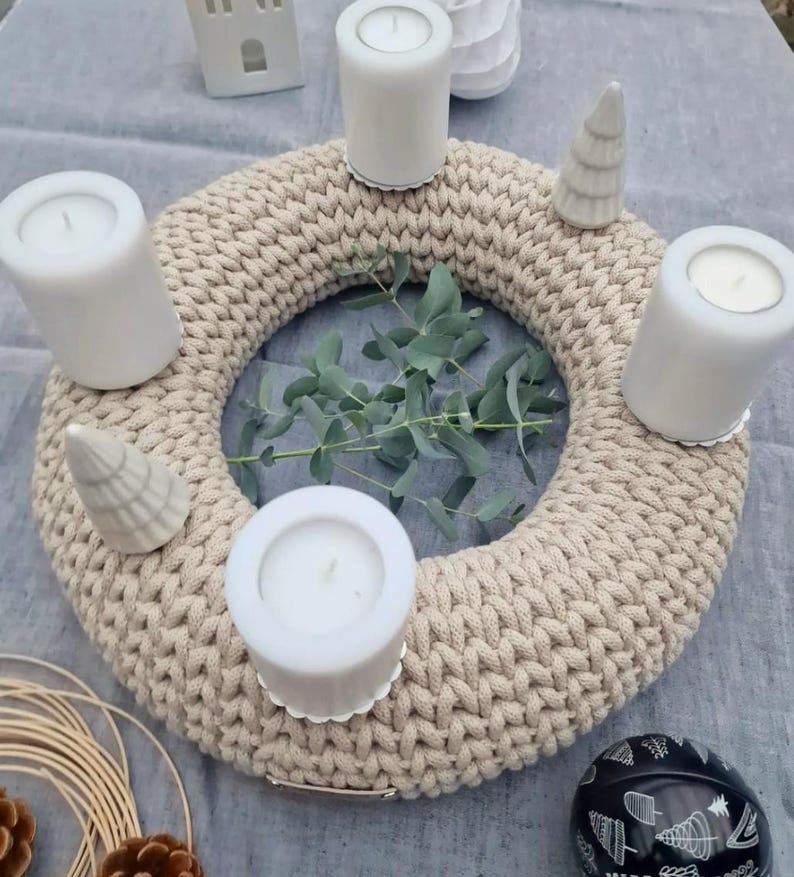 Puede incluir: Una corona de punto grueso beige con cuatro velas blancas y dos peque&ntilde;as figuras de &aacute;rboles de Navidad de cer&aacute;mica. Ramas de eucalipto est&aacute;n en el centro. Una bola decorativa en blanco y negro y otras decoraciones navide&ntilde;as est&aacute;n en el fondo.