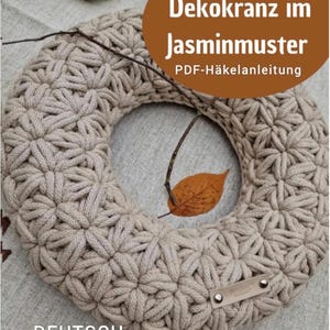 Könnte beinhalten: Ein beigefarbener Häkelkranz mit einem sich wiederholenden sternförmigen Jasminmuster. Unten rechts ist ein kleines rechteckiges Etikett in Lederoptik mit zwei Metallnieten befestigt. Der Text auf dem Bild lautet: Dekokranz im Jasminmuster PDF-Häkelanleitung.