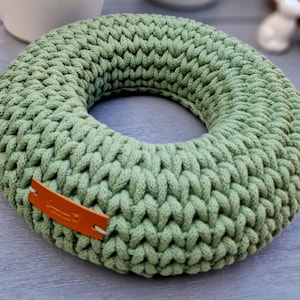 Puede incluir: Una corona circular de ganchillo verde salvia. La corona est&aacute; hecha de hilo grueso y tiene una textura trenzada. Una peque&ntilde;a etiqueta de cuero marr&oacute;n est&aacute; adherida a la corona. La corona es un art&iacute;culo decorativo.