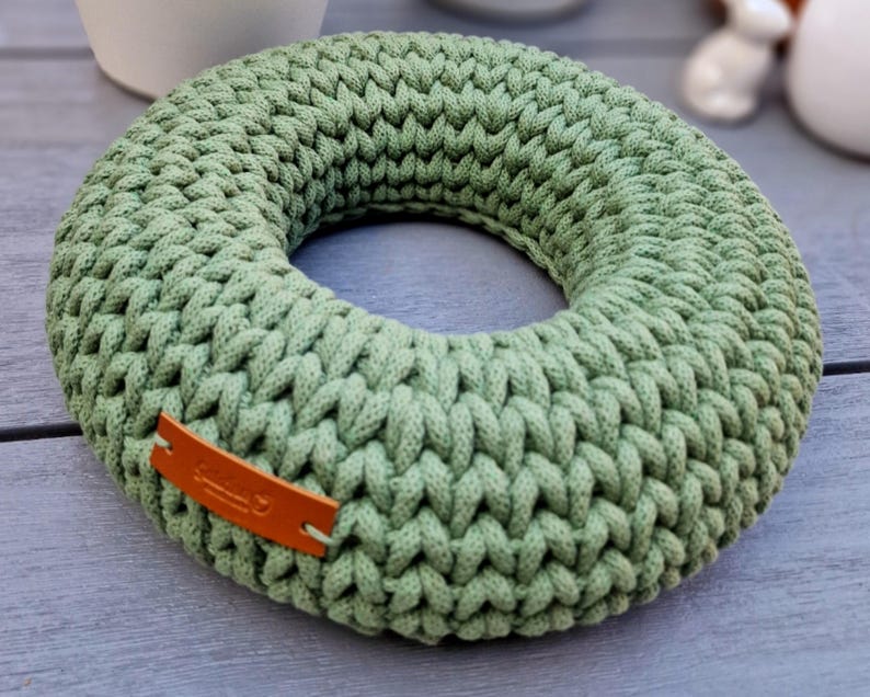 Puede incluir: Una corona circular de ganchillo verde salvia. La corona est&aacute; hecha de hilo grueso y tiene una textura trenzada. Una peque&ntilde;a etiqueta de cuero marr&oacute;n est&aacute; adherida a la corona. La corona es un art&iacute;culo decorativo.