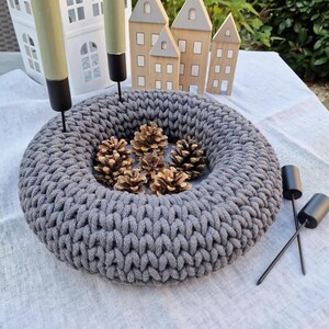 Puede incluir: Una corona gris tejida con un dise&ntilde;o circular, llena de varios conos de pino. La corona est&aacute; rodeada por dos candelabros negros con velas verdes. En el fondo hay casas decorativas de madera y una casa decorativa blanca.