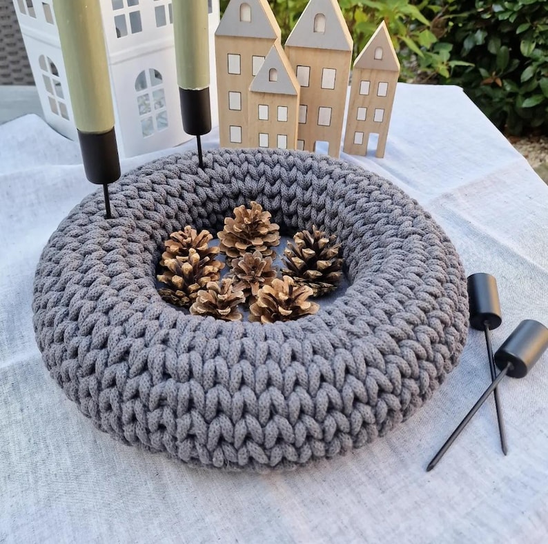 Puede incluir: Una corona gris tejida con un dise&ntilde;o circular, llena de varios conos de pino. La corona est&aacute; rodeada por dos candelabros negros con velas verdes. En el fondo hay casas decorativas de madera y una casa decorativa blanca.