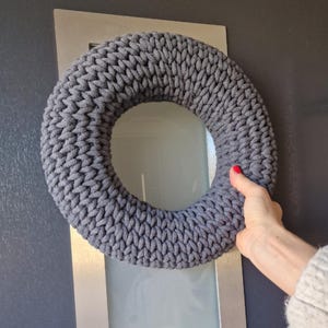 Puede incluir: Una corona gris tejida con una apariencia gruesa y texturizada. La corona circular parece estar hecha a mano. El color es un gris apagado, adecuado para varios estilos de decoraci&oacute;n del hogar.