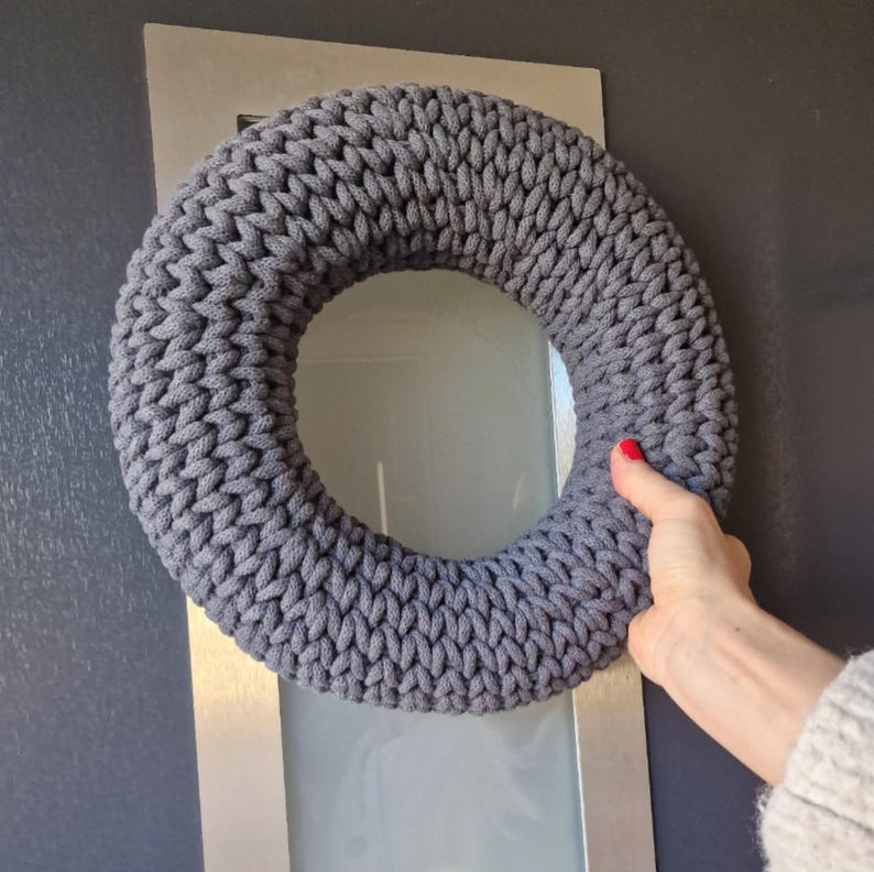 Puede incluir: Una corona gris tejida con una apariencia gruesa y texturizada. La corona circular parece estar hecha a mano. El color es un gris apagado, adecuado para varios estilos de decoraci&oacute;n del hogar.