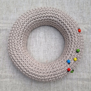 Puede incluir: Una corona de punto beige con un dise&ntilde;o circular. La corona est&aacute; decorada con peque&ntilde;os adornos de mariquitas de colores. La corona se exhibe sobre una superficie texturizada de color neutro.