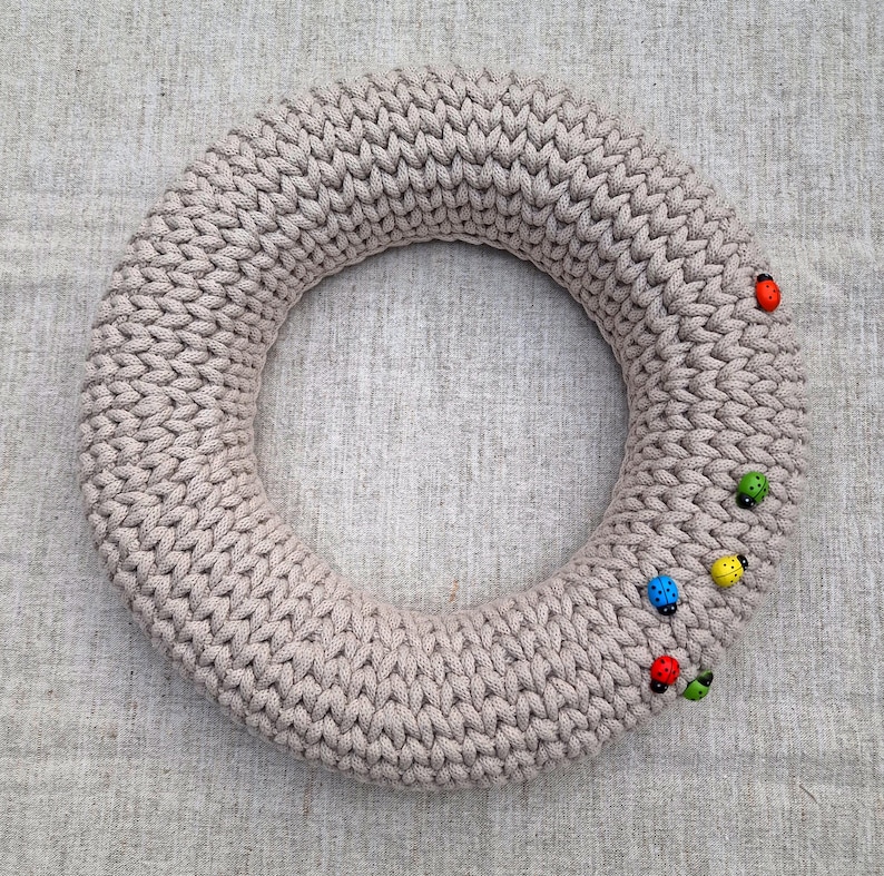Puede incluir: Una corona de punto beige con un dise&ntilde;o circular. La corona est&aacute; decorada con peque&ntilde;os adornos de mariquitas de colores. La corona se exhibe sobre una superficie texturizada de color neutro.