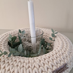 Puede incluir: Un portavelas de ganchillo color crema con una vela de cristal que contiene una vela blanca y musgo decorativo. Ramas de eucalipto se colocan alrededor del cristal. Una peque&ntilde;a etiqueta marr&oacute;n con el texto "Galana's handmade" est&aacute; adjunta.