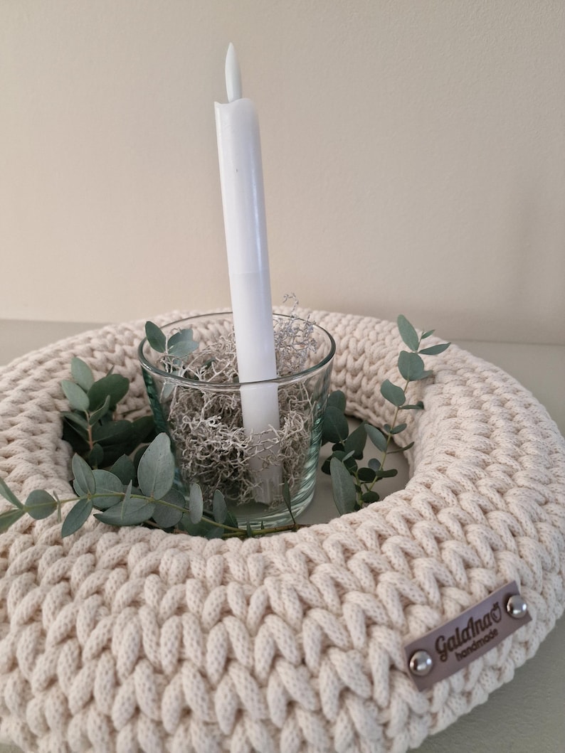 Puede incluir: Un portavelas de ganchillo color crema con una vela de cristal que contiene una vela blanca y musgo decorativo. Ramas de eucalipto se colocan alrededor del cristal. Una peque&ntilde;a etiqueta marr&oacute;n con el texto "Galana's handmade" est&aacute; adjunta.