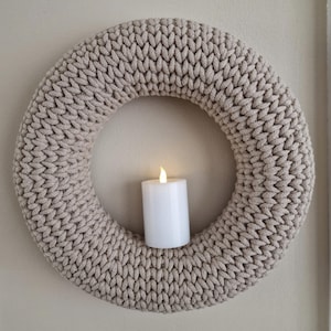 Puede incluir: Una corona beige tejida con un dise&ntilde;o grueso y texturizado. Una vela cil&iacute;ndrica blanca con una llama parpadeante est&aacute; centrada dentro de la corona. La corona se exhibe contra una pared de color neutro, creando un acento decorativo y acogedor.