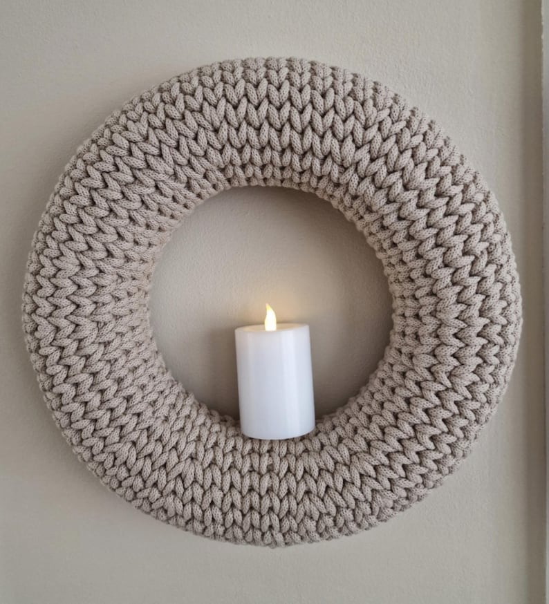 Puede incluir: Una corona beige tejida con un dise&ntilde;o grueso y texturizado. Una vela cil&iacute;ndrica blanca con una llama parpadeante est&aacute; centrada dentro de la corona. La corona se exhibe contra una pared de color neutro, creando un acento decorativo y acogedor.