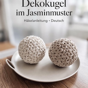 May include: Two spherical crochet items featuring a jasmine stitch pattern in light beige and tan yarn. The text above reads: Dekokugel im Jasminmuster Häkelanleitung • Deutsch. These round, textured objects are displayed on a smooth, light-coloured plate.