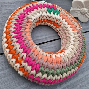 Puede incluir: Una colorida corona de ganchillo hecha a mano con un dise&ntilde;o circular. La corona presenta una variedad de colores, incluyendo rosa, naranja, verde y crema. El hilo est&aacute; tejido apretadamente, creando una superficie texturizada. La corona se utiliza probablemente para la decoraci&oacute;n del hogar.