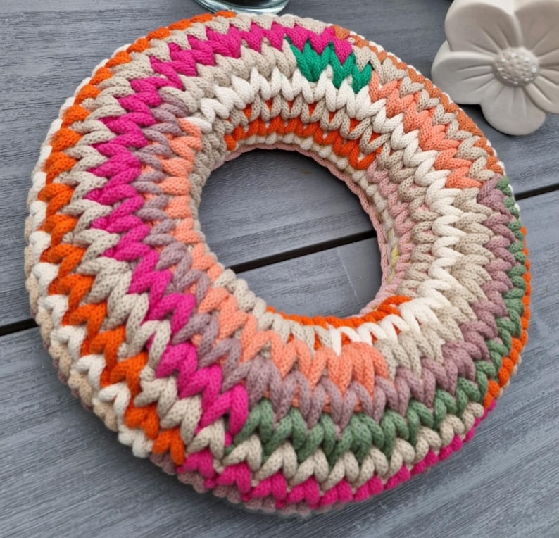 Puede incluir: Una colorida corona de ganchillo hecha a mano con un dise&ntilde;o circular. La corona presenta una variedad de colores, incluyendo rosa, naranja, verde y crema. El hilo est&aacute; tejido apretadamente, creando una superficie texturizada. La corona se utiliza probablemente para la decoraci&oacute;n del hogar.
