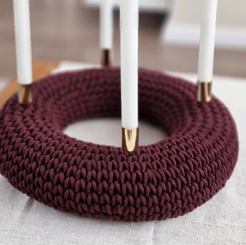 Puede incluir: Un portavelas de ganchillo burdeos con cuatro velas blancas. El portavelas tiene un dise&ntilde;o circular con detalles dorados en la base de cada vela. El soporte est&aacute; hecho de un hilo grueso y texturizado.