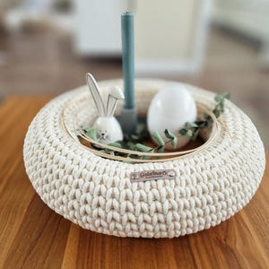 Puede incluir: Centro de mesa de Pascua de ganchillo color crema con dise&ntilde;o circular. Presenta un conejito de cer&aacute;mica blanca, un huevo blanco, una vela verde azulado y vegetaci&oacute;n. El centro de mesa est&aacute; sobre una superficie de madera.