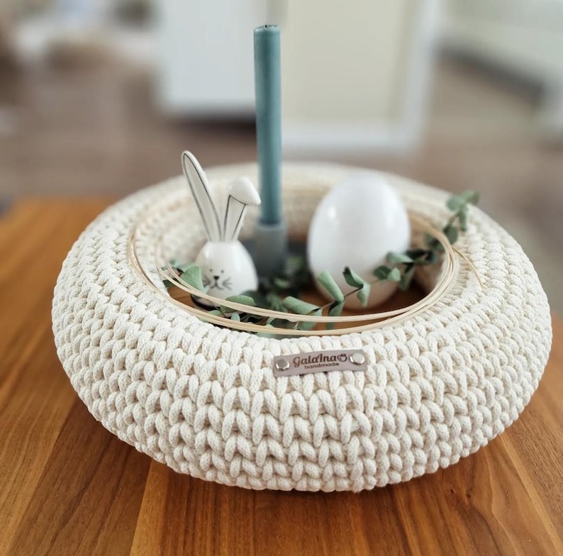 Puede incluir: Centro de mesa de Pascua de ganchillo color crema con dise&ntilde;o circular. Presenta un conejito de cer&aacute;mica blanca, un huevo blanco, una vela verde azulado y vegetaci&oacute;n. El centro de mesa est&aacute; sobre una superficie de madera.