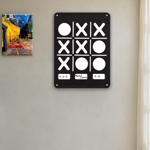 May include: Black tic-tac-toe game board with white X's and O's. The board is rectangular with rounded corners and is mounted on a light gray wall. The text "+++" and "mors/sora" is printed on the bottom of the board.