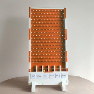 Pode incluir: Um jogo de Plinko de madeira com uma superfície de jogo laranja e pinos brancos. A moldura é branca com listras laranja e brancas. O jogo tem uma série de ranhuras na parte inferior com bolas coloridas e etiquetas com nomes. Os nomes nas etiquetas são: Jack, Maya, Walt, Ana, Mory e Kate.