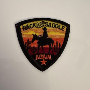 May include: Embroidered patch with a shield shape. The patch features a silhouette of a cowboy on a horse against a sunset backdrop of orange, yellow, and red. Text reads "BACK IN THE SADDLE AGAIN" with a star.