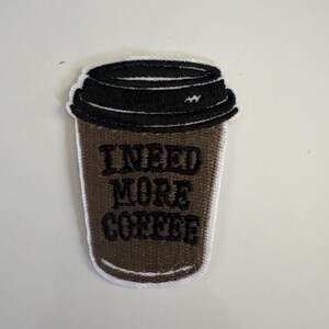 May include: Embroidered patch of a coffee cup with a black lid. The brown cup features the words "I NEED MORE COFFEE" in black. The patch has a white border, suitable for sewing onto clothing or bags.