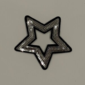 May include: A silver sequined star-shaped patch with a black border. The sequins are densely packed, creating a shimmering effect. The patch is likely for decorative purposes, such as embellishing clothing or accessories.