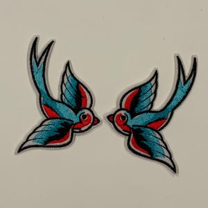 May include: Two embroidered patches featuring stylized swallow birds. The birds have teal blue wings with red accents and black outlines. The patches are on a white background.