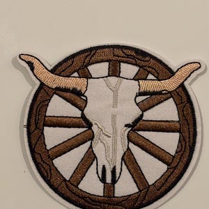 Puede incluir: Parche bordado con una calavera de toro blanca con cuernos beige, centrada en un diseño de rueda de carro marrón. El parche tiene un borde negro y es probablemente para aplicar en ropa o accesorios. Temática del oeste.