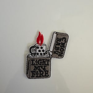 May include: Embroidered patch featuring a brown lighter with a red flame. The lighter has the text "LIGHT MY FIRE" and "NON BABI" on it. The patch is on a white background.