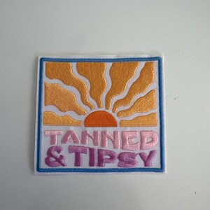 Puede incluir: Parche bordado de forma cuadrada, con un diseño de sol naranja estilizado con rayos y las palabras "TANNED & TIPSY" en rosa y morado. El parche tiene un fondo blanco con un borde azul.