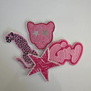 May include: A collection of embroidered patches in shades of pink. Designs include a pink leopard, a pink panther head with star-shaped eyes, a pink star with zebra stripes, and the word "Girl" in cursive.