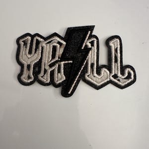 May include: Embroidered patch with the word "Y'ALL" in a stylized font, featuring a black glitter lightning bolt. The letters have a white outline and a leopard print pattern.