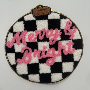 May include: A round Christmas ornament with a black and white checkered pattern. The words "Merry & Bright" are in pink, textured lettering. The ornament has a brown top and a gold border.