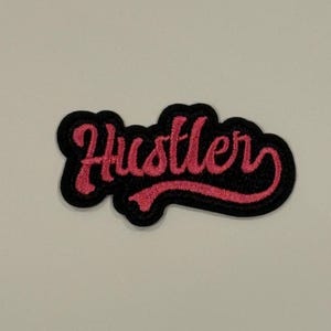 Könnte beinhalten: Gestickter Aufnäher mit dem Wort "Hustler" in rosa Schrift. Der Text ist rosa umrandet und vor einem schwarzen Hintergrund platziert. Der Aufnäher hat eine gebogene rechteckige Form.
