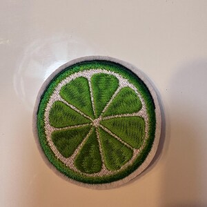 May include: Embroidered patch featuring a lime slice design. The patch has a bright green center with white and gray accents, and a gray border. The design is circular and detailed, resembling a realistic citrus fruit.