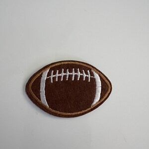 May include: A brown football-shaped patch with white stitching and a brown border. The patch is an oval shape with white stitching across the center, resembling the laces of a football. The patch is likely for decorative purposes.
