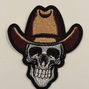Puede incluir: Parche bordado con una calavera que lleva un sombrero de vaquero. El sombrero es marrón y beige, con un borde negro. La calavera es plateada y negra, con dientes visibles. El parche tiene un borde negro.