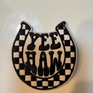 May include: Embroidered patch in the shape of a horseshoe with the words "YEE HAW" in black block letters. The horseshoe is white with a black and white checkered pattern on the edges.