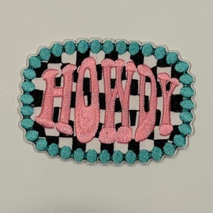 May include: Pink embroidered patch with the word "HOWDY" in a bubble font. The patch has a black and white checkered background and a teal beaded border. The patch is oval-shaped.