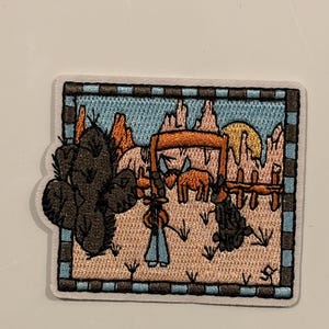 Peut inclure: Écusson brodé représentant une scène de désert avec un cactus, un cow-boy et un chien. L'écusson a une forme rectangulaire avec une bordure bleue et noire. Le fond représente un coucher de soleil sur le désert.
