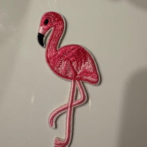 Peut inclure: Écusson brodé représentant un flamant rose. Le flamant rose est rose vif avec un bec et un œil noirs. Il est représenté sur une patte, le cou courbé.