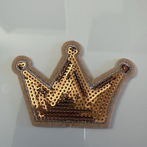 May include: A gold sequined crown applique with a tan felt backing and silver trim. The crown has three points and is designed for embellishment on clothing or accessories. The sequins are arranged in rows, creating a shimmering effect.