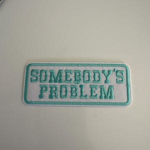 May include: White rectangular patch with a teal border and teal embroidered text that reads "SOMEBODY'S PROBLEM". The patch is likely designed to be sewn or ironed onto clothing or accessories.