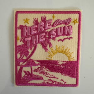 May include: Embroidered patch with the text "Here Comes The Sun" in pink, featuring a beach scene with palm trees, a sunset, and waves. The patch has a cream background and a pink border.