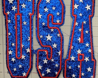 May include: Embroidered patches spelling out "USA" in a bold, block letter style. The letters are blue with a glittery texture, outlined in red, and filled with white stars. A patriotic theme.